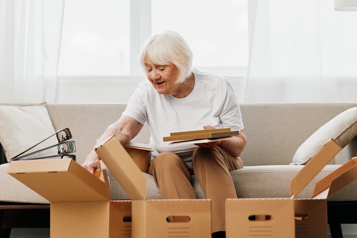 Assisted Living in Sarasota elderly woman sits on a sofa at home with boxes. collecting things with memories albums with photos and photo frames moving to a new place cleaning things and a happy smile. Lifestyle retirement.